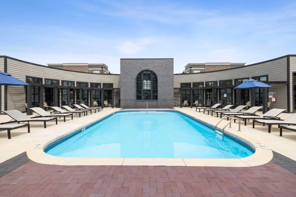Outdoor swimming pool with lounge chairs and blue umbrellas surrounding it, set in front of a modern building with large windows under a clear sky.