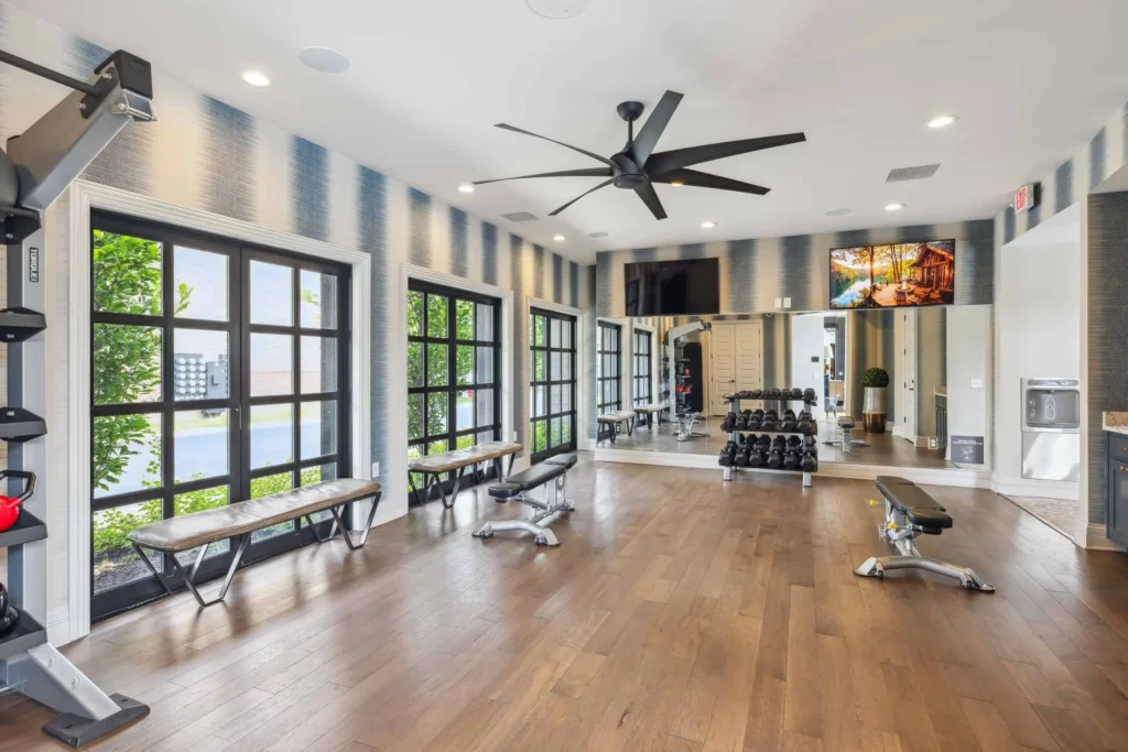 Modern gym with wood flooring, free weights, benches, large mirrors, wall-mounted TV, and floor-to-ceiling windows allowing natural light.