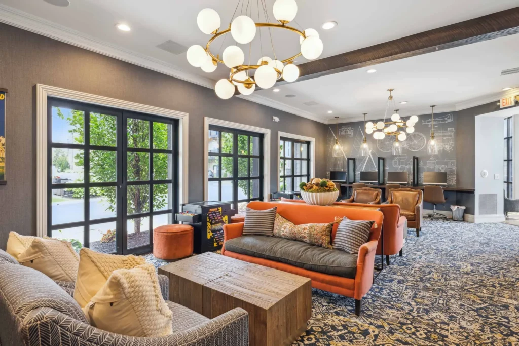 Modern lounge with patterned carpet, gray and orange seating, wooden coffee table, large windows, and spherical pendant lights. Desks and chalkboard wall are visible in the background.
