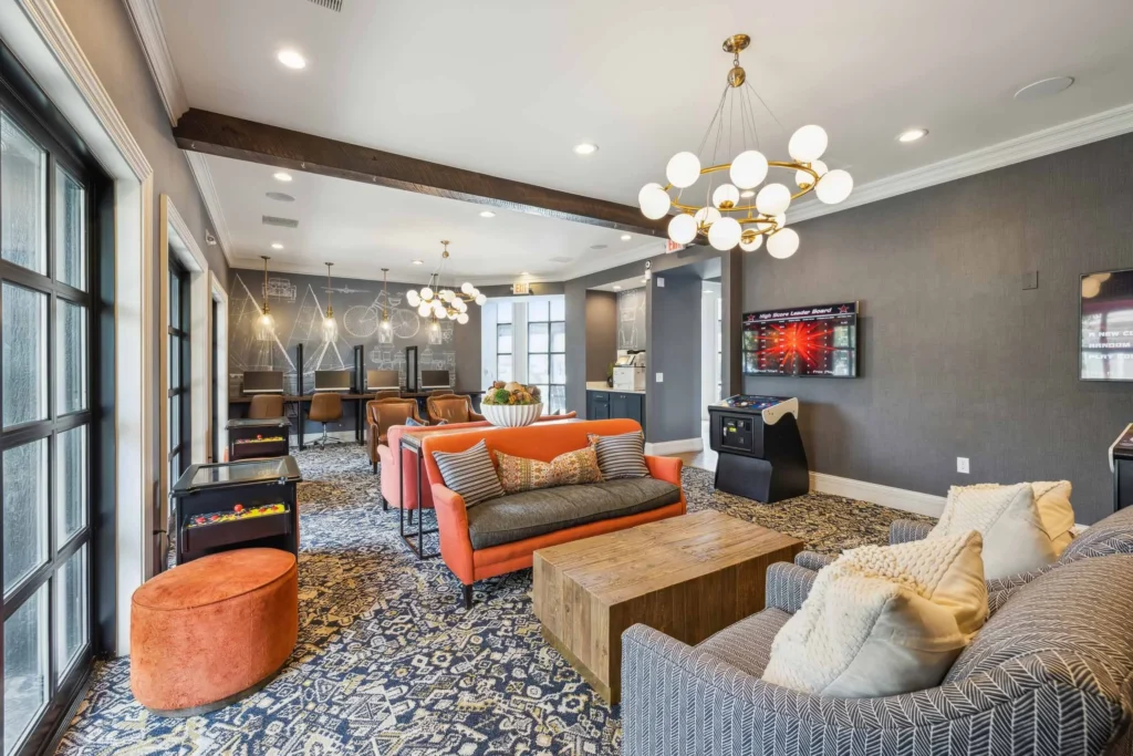 Modern lounge area with patterned carpet, orange sofa, gray armchairs, wooden coffee table, TV, arcade game, bar seating, and large windows.