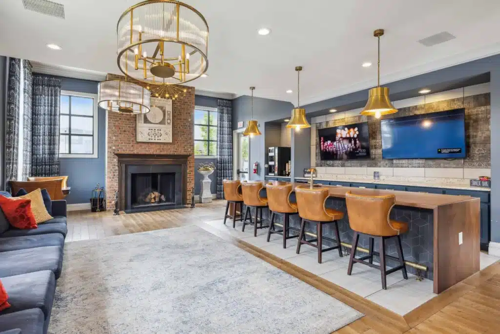 Modern lounge with leather bar stools at a kitchen counter, a brick fireplace, wall-mounted TV, and gold pendant lights, featuring hardwood floors and large windows.