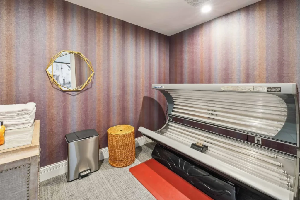 A tanning bed is positioned against a striped wallpapered wall, next to a mirror, a trash can, a wicker basket, and a stack of towels.