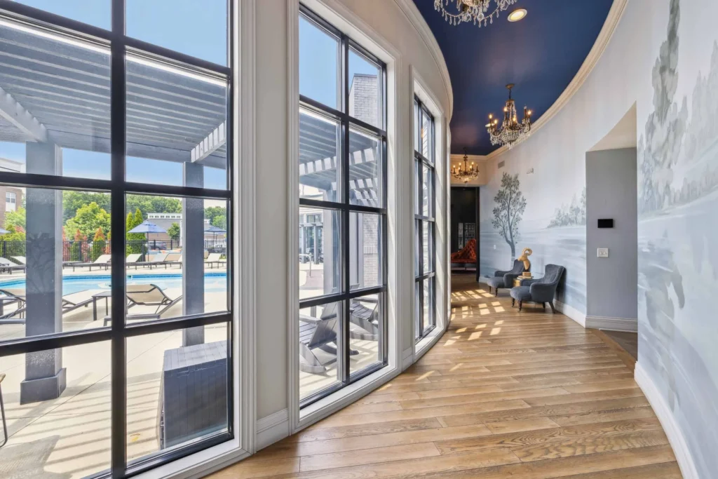 Curved hallway with large floor-to-ceiling windows overlooking a pool area, wood flooring, chandeliers, and wall art depicting trees.