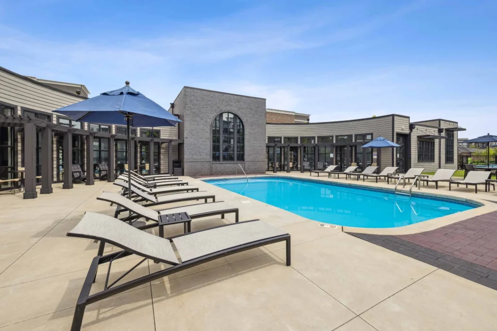Outdoor pool area with lounge chairs, umbrellas, and a modern building in the background under a clear blue sky.