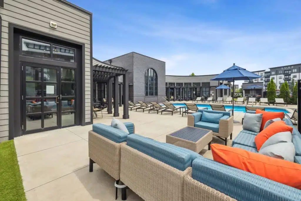 Outdoor lounge area with wicker furniture and colorful cushions next to a swimming pool, surrounded by modern apartment buildings under a blue sky.