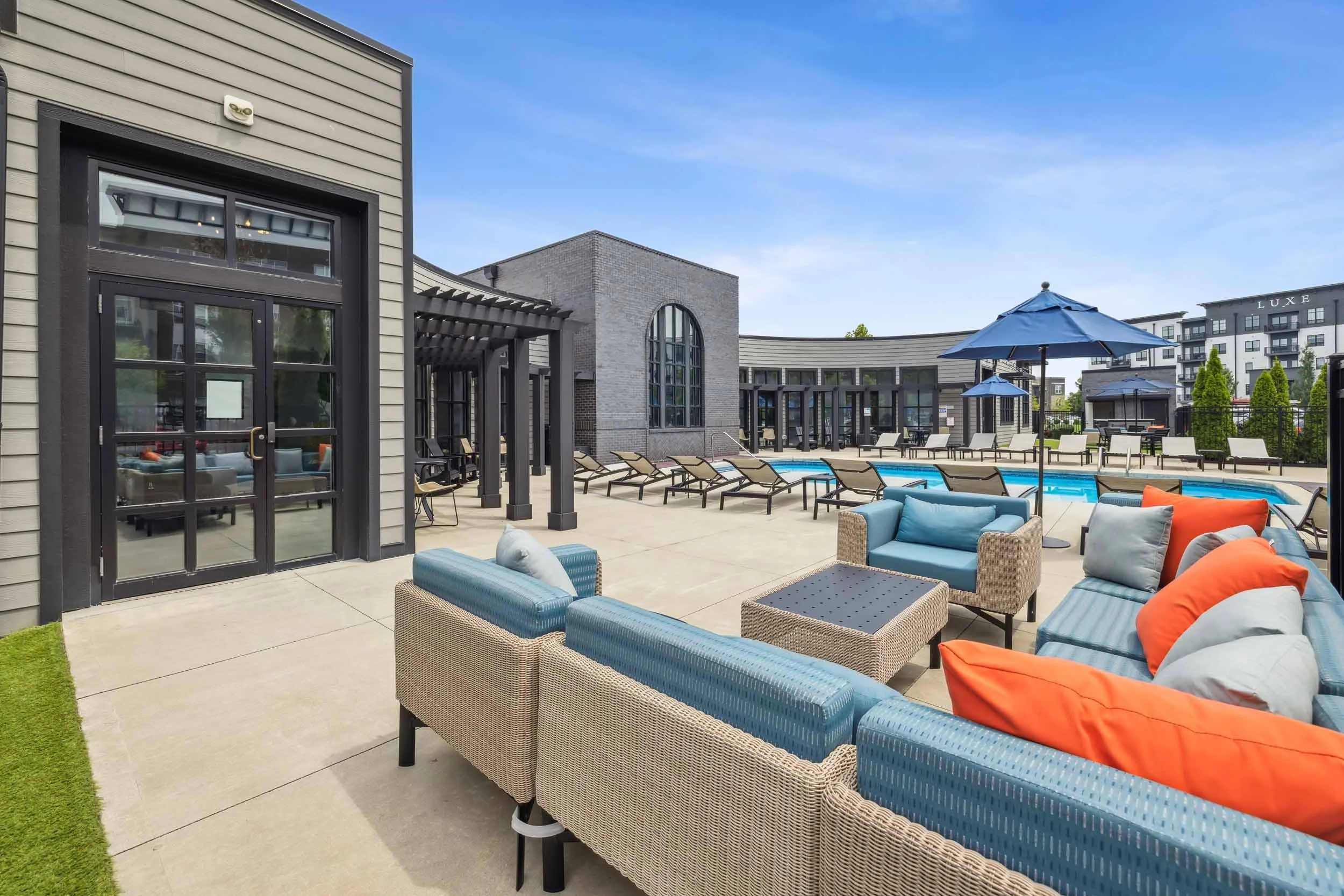 Outdoor lounge area with wicker furniture and colorful cushions next to a swimming pool, surrounded by modern apartment buildings under a blue sky.