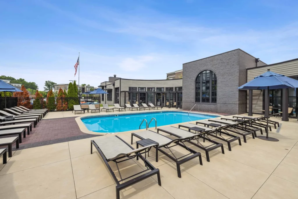 Outdoor swimming pool with lounge chairs and umbrellas on a sunny day, adjacent to a modern building with large arched windows.