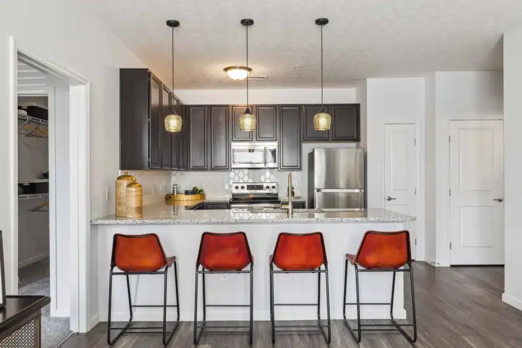 Modern kitchen with dark cabinets, stainless steel appliances, granite countertops, pendant lights, and four orange barstools at a breakfast bar. Closet and door visible in the background.