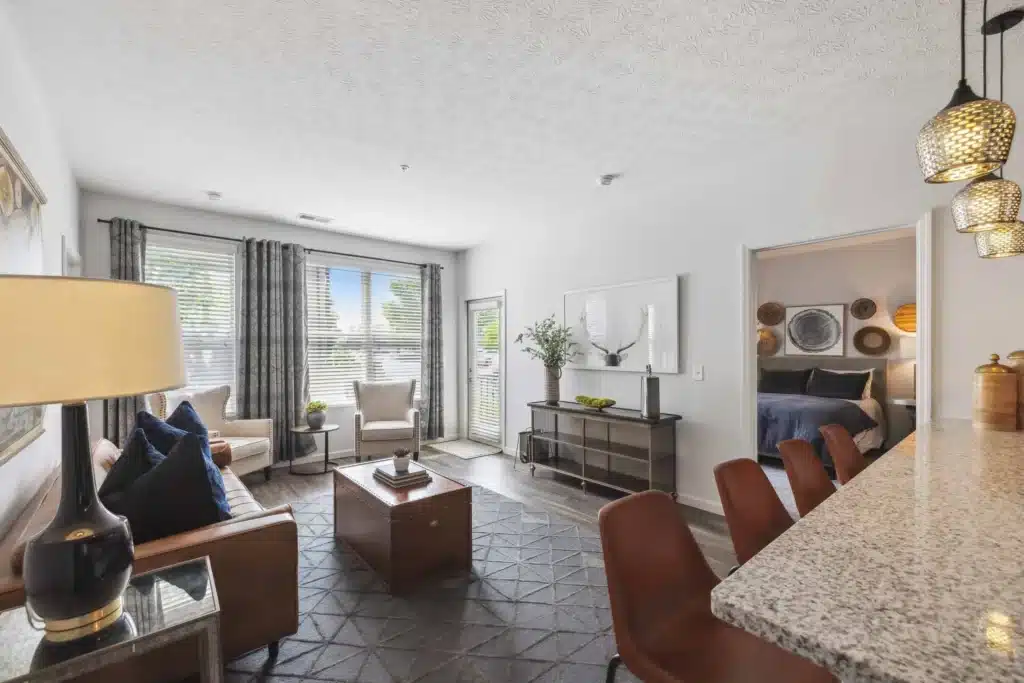 Modern living room with a brown sofa, two armchairs, a coffee table, and large windows; open kitchen with granite countertop and pendant lights, bedroom visible through doorway.