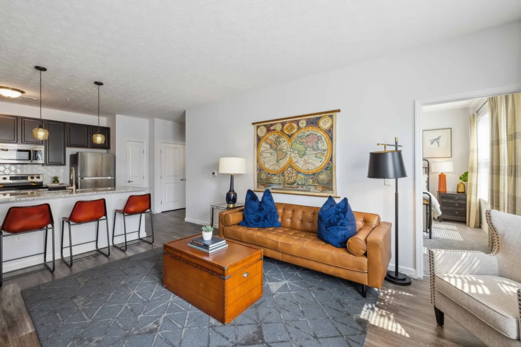 Modern living room with a brown leather sofa, wall map art, orange bar stools at a kitchen counter, and a chest-style coffee table on a blue rug. Bedroom visible through an open door.