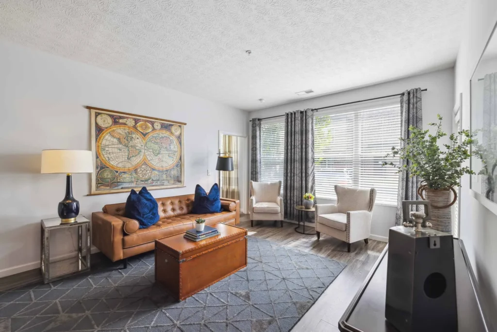 Bright living room with a brown leather sofa, two white armchairs, large map artwork, and a trunk coffee table on a patterned rug near a window with blinds and curtains.