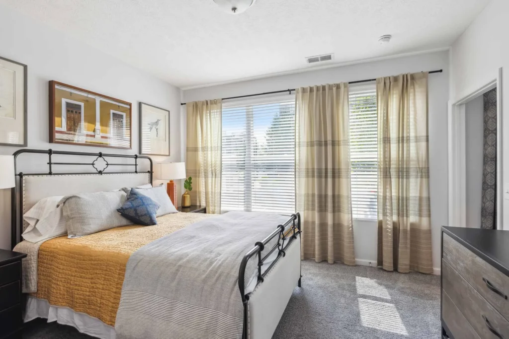 Modern bedroom with a metal-frame bed, yellow and gray bedding, framed art on white walls, a dresser, and large windows with sheer curtains letting in natural light.