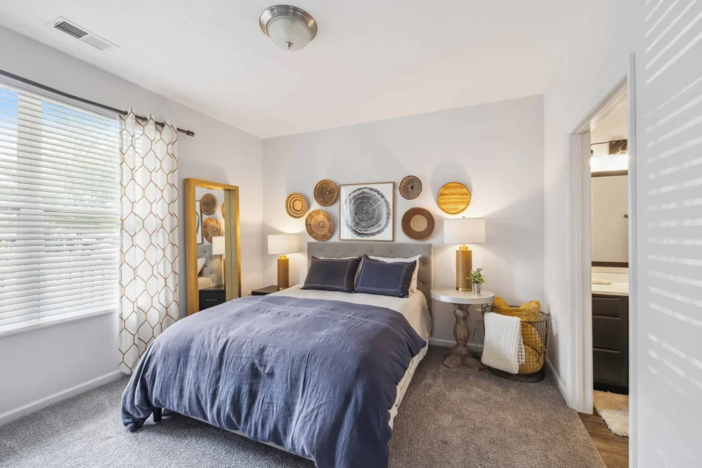A bright bedroom with a double bed, dark blue bedding, decorative wall baskets, a tall mirror, side tables with lamps, a window with patterned curtains, and an open door to a bathroom.