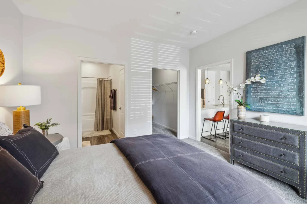 Modern bedroom with a large bed, dresser, wall art, en-suite bathroom, walk-in closet, and a desk with an orange chair. Natural light streams in through blinds.