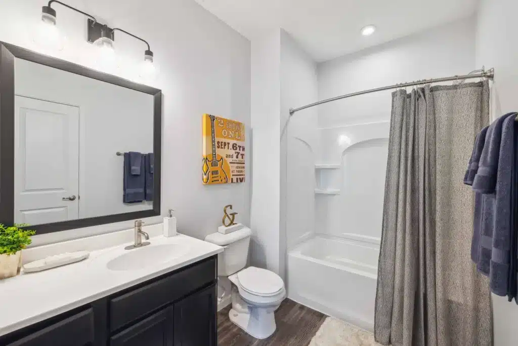 Modern bathroom with a white bathtub and shower, gray curtain, toilet, dark vanity with sink, wall mirror, towels, and decorative items.