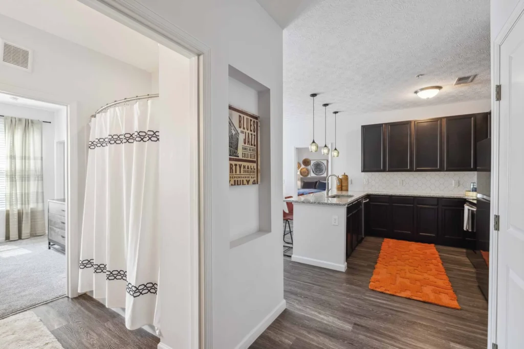 View of a hallway leading to a bathroom with a shower curtain on the left and a modern kitchen with dark cabinets, pendant lights, and an orange rug on the right.