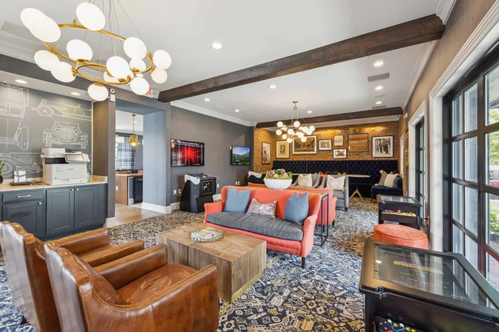 Modern lounge with leather and fabric seating, a wooden coffee table, wall art, patterned carpet, arcade machines, and large windows letting in natural light.
