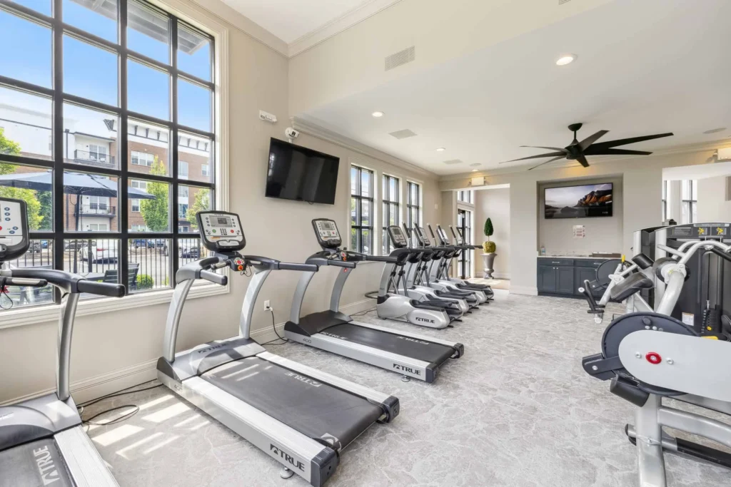 A modern fitness center with treadmills, ellipticals, and stationary bikes facing large windows and a wall-mounted TV, with bright natural light filling the room.