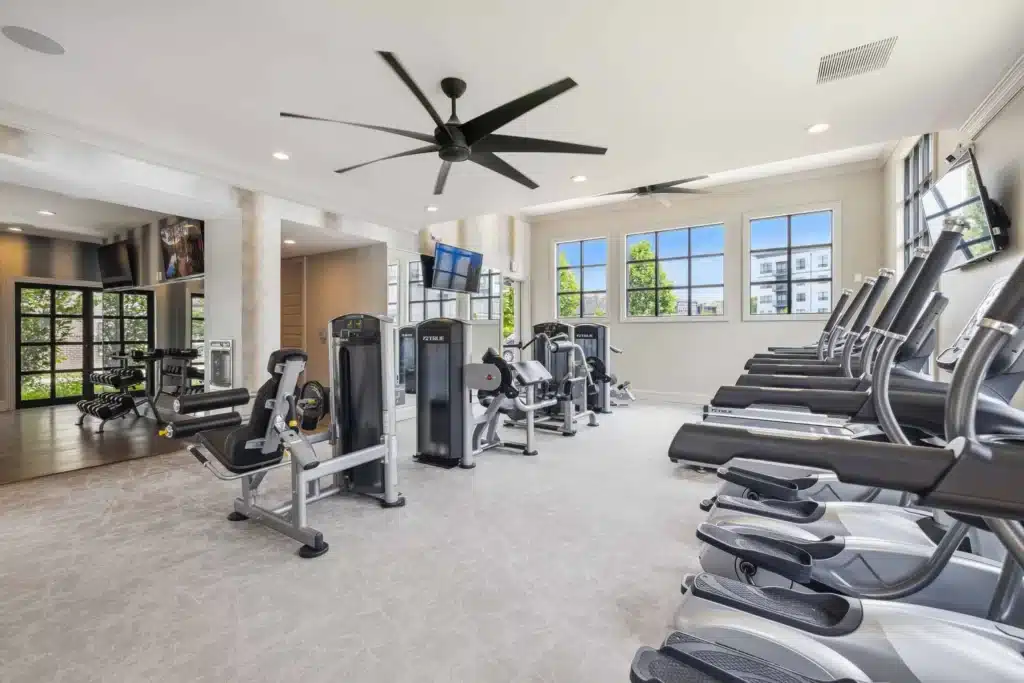 Bright, modern gym with treadmills, stationary bikes, weight machines, large windows, TV screens, and ceiling fans.