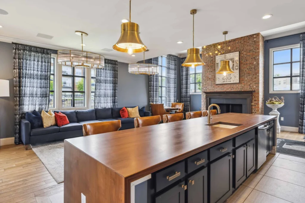 Modern open-plan living room and kitchen with a large wooden island, dark cabinetry, leather sofa, brick fireplace, large windows, and gold pendant lighting.