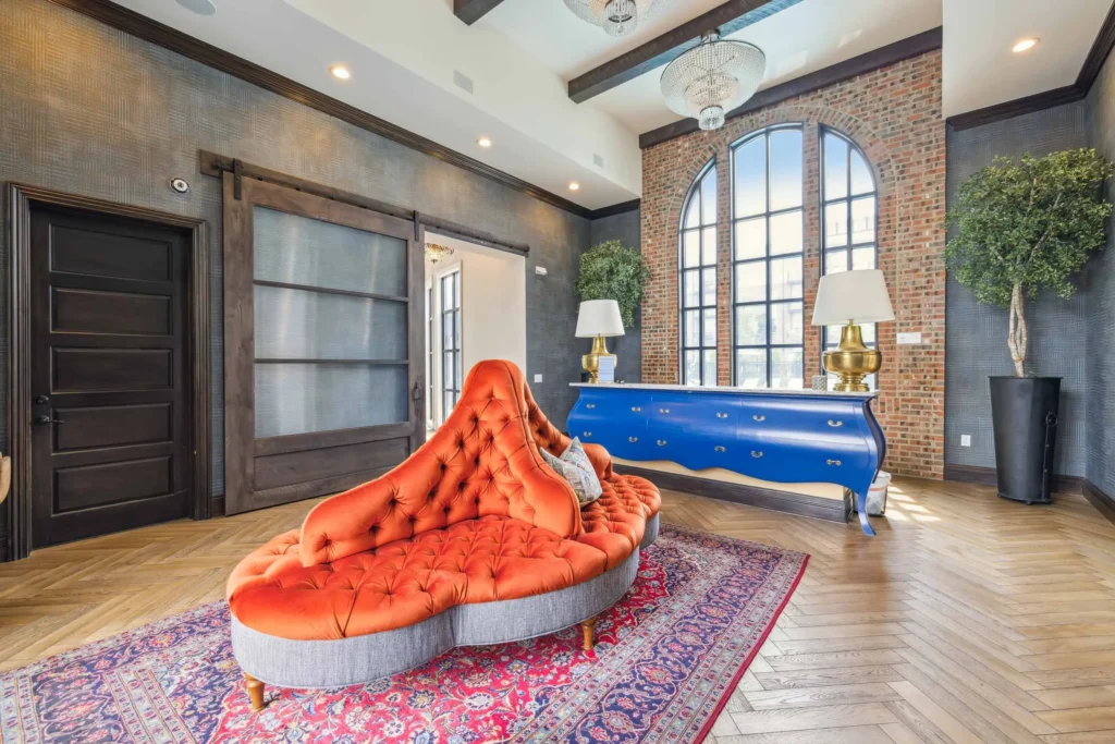 Modern living room with a red tufted sofa, patterned rug, large arched window, blue dresser, black doors, and potted trees on a herringbone wood floor.