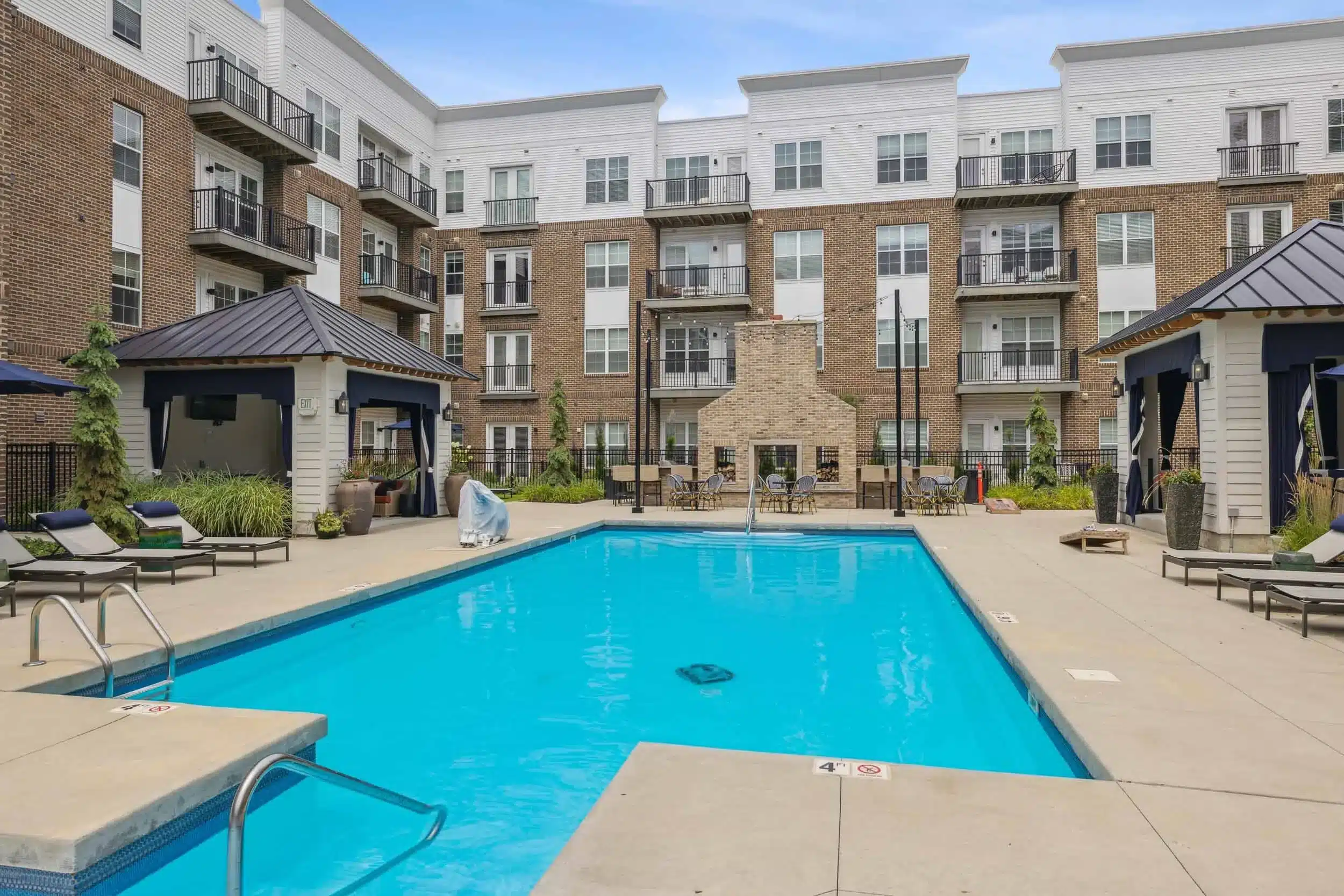Outdoor swimming pool surrounded by lounge chairs, cabanas, and a four-story apartment building with balconies in the background.