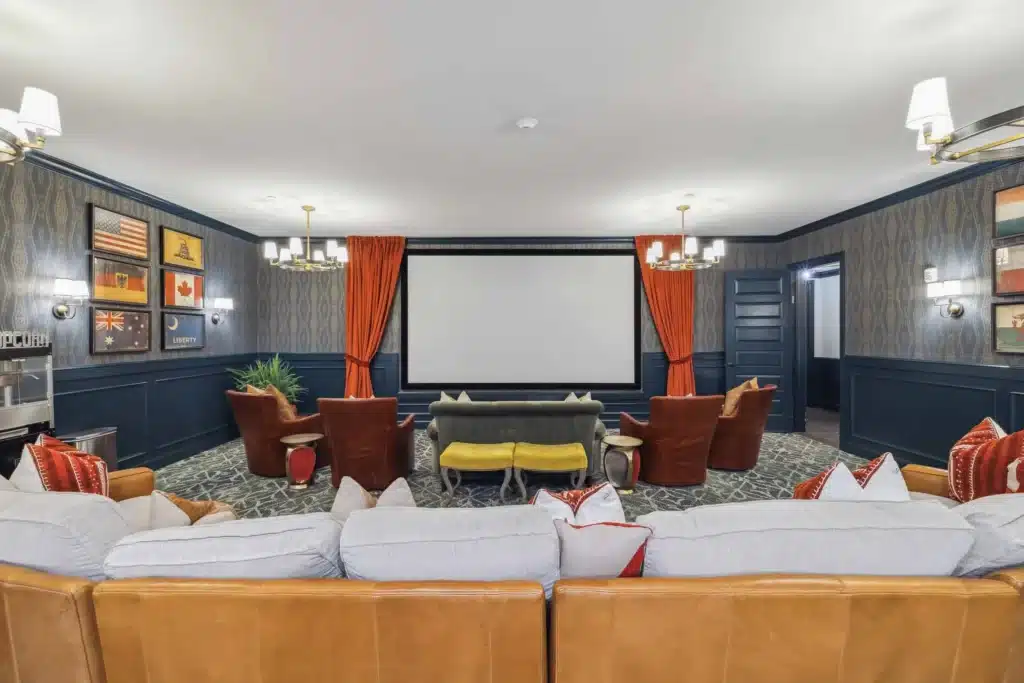 Home theater room with a large projector screen, sofas and armchairs, patterned carpet, wall sconces, and framed flags on dark blue paneled walls.