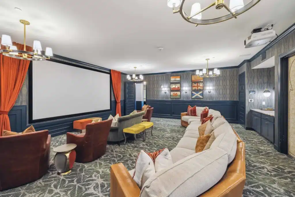 A home theater room with a large projector screen, various upholstered seating, patterned carpet, wall sconces, and decorative accents.