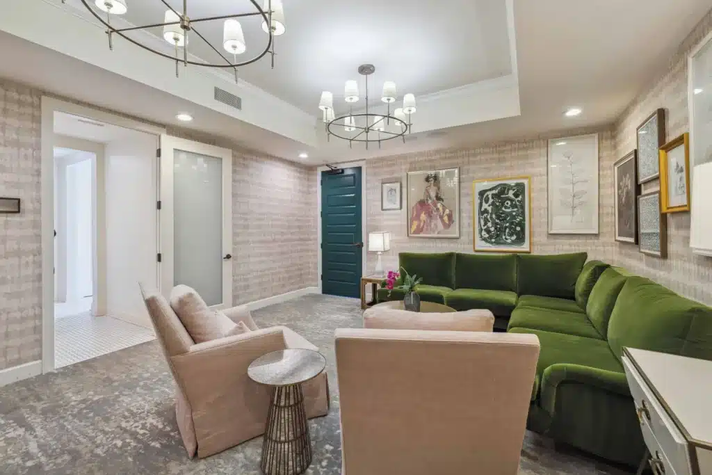 A modern living room features a green sectional sofa, two beige armchairs, framed artwork on the walls, and two ceiling light fixtures.