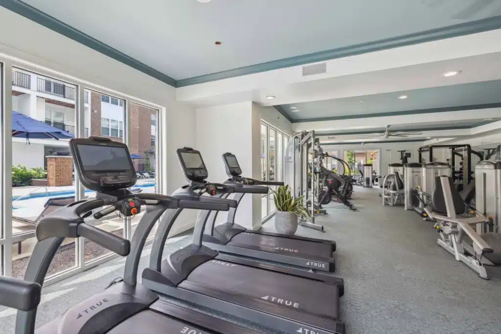 Modern gym with treadmills, weight machines, and stationary bikes next to large windows overlooking an outdoor pool area.