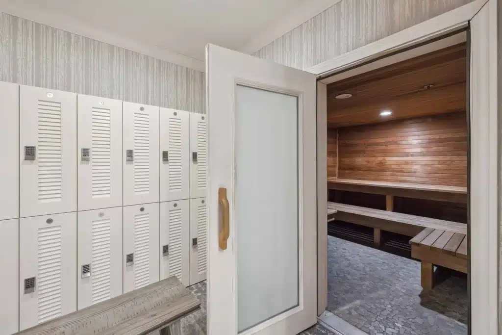 Locker room with white lockers and a wooden bench, showing an open door leading to a wood-paneled sauna with benches inside.
