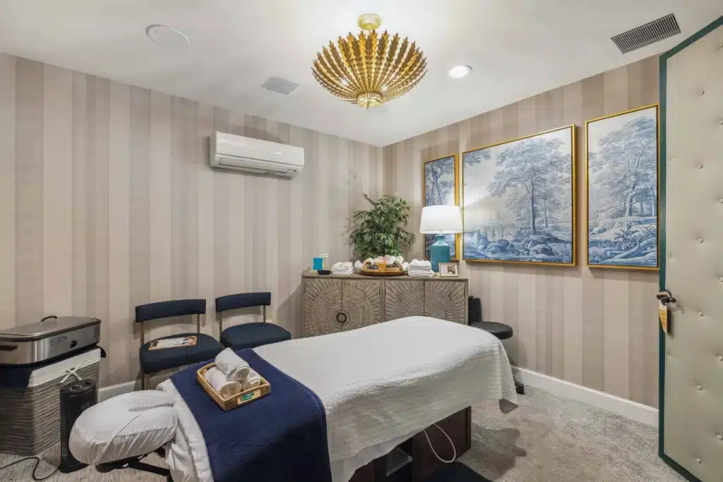 A neatly arranged massage room with a massage table, chairs, decorative artwork, a lamp, and a gold chandelier under soft lighting.