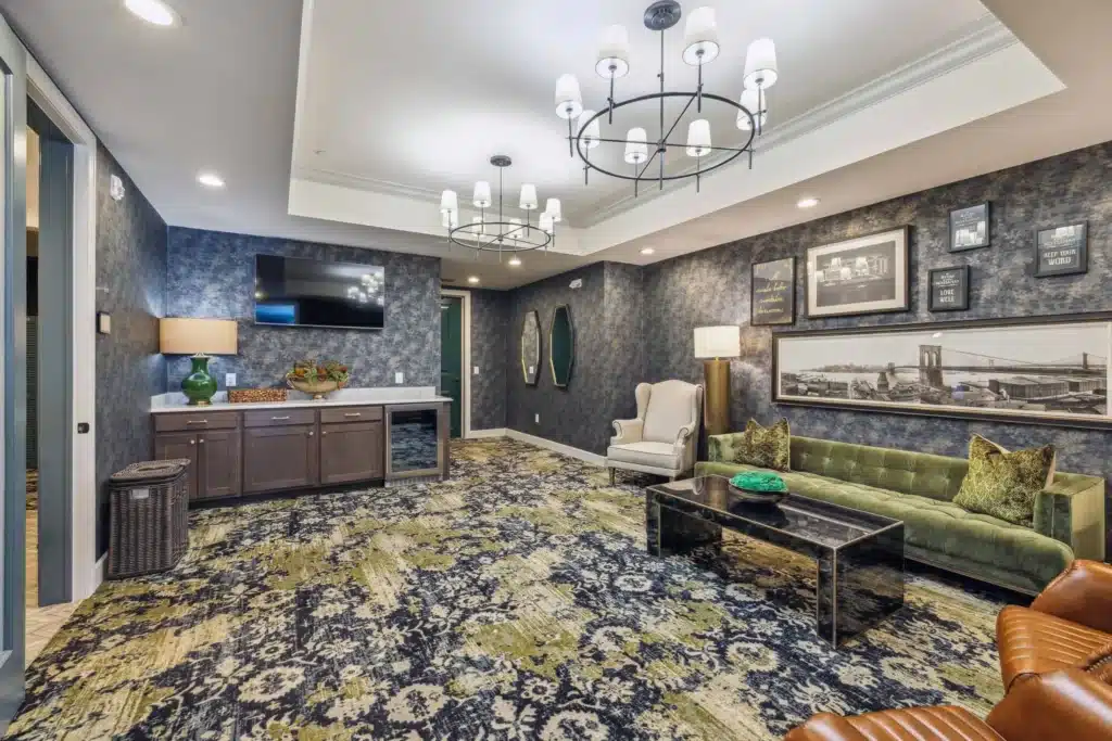 A modern lounge with a patterned carpet, green sofa, wingback chair, wall art, two chandeliers, a TV, and a cabinet with plants and lamps.