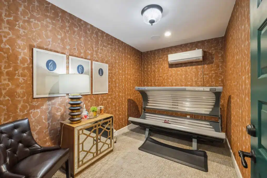 A small room with patterned brown wallpaper, a tanning bed, a leather chair, a mirrored cabinet with a lamp, and two framed pictures on the wall.