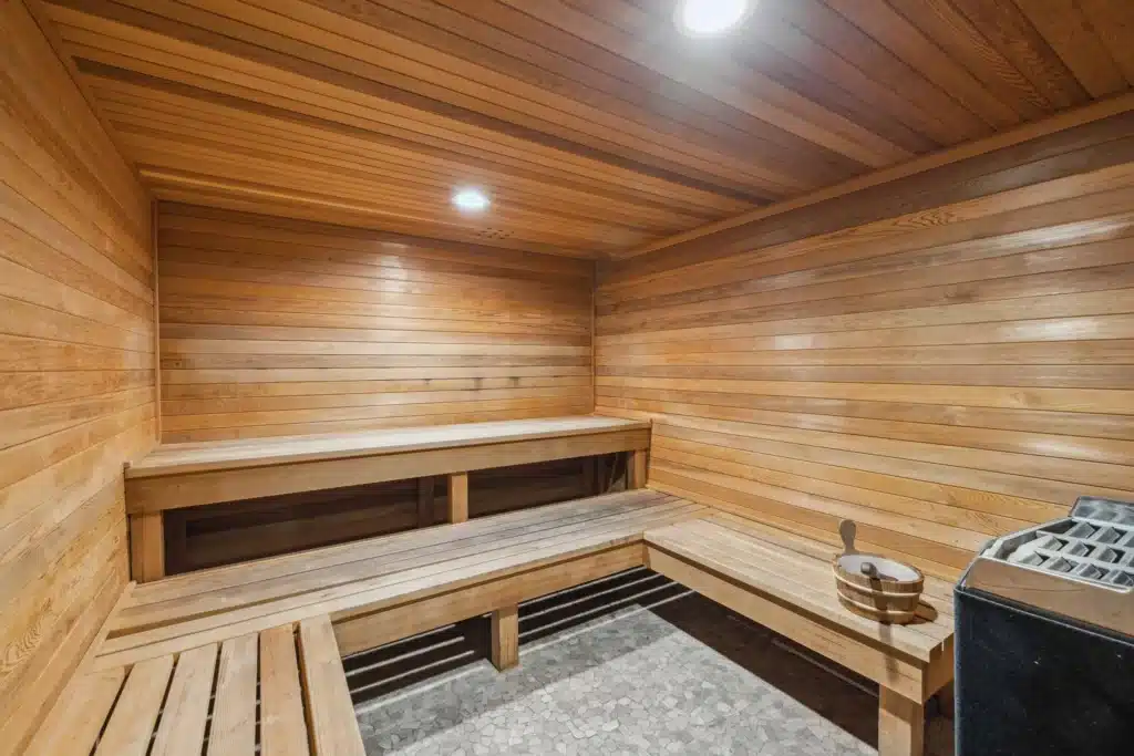 Interior of a wooden sauna with tiered bench seating, overhead lights, a bucket with ladle, and a sauna heater in the corner.