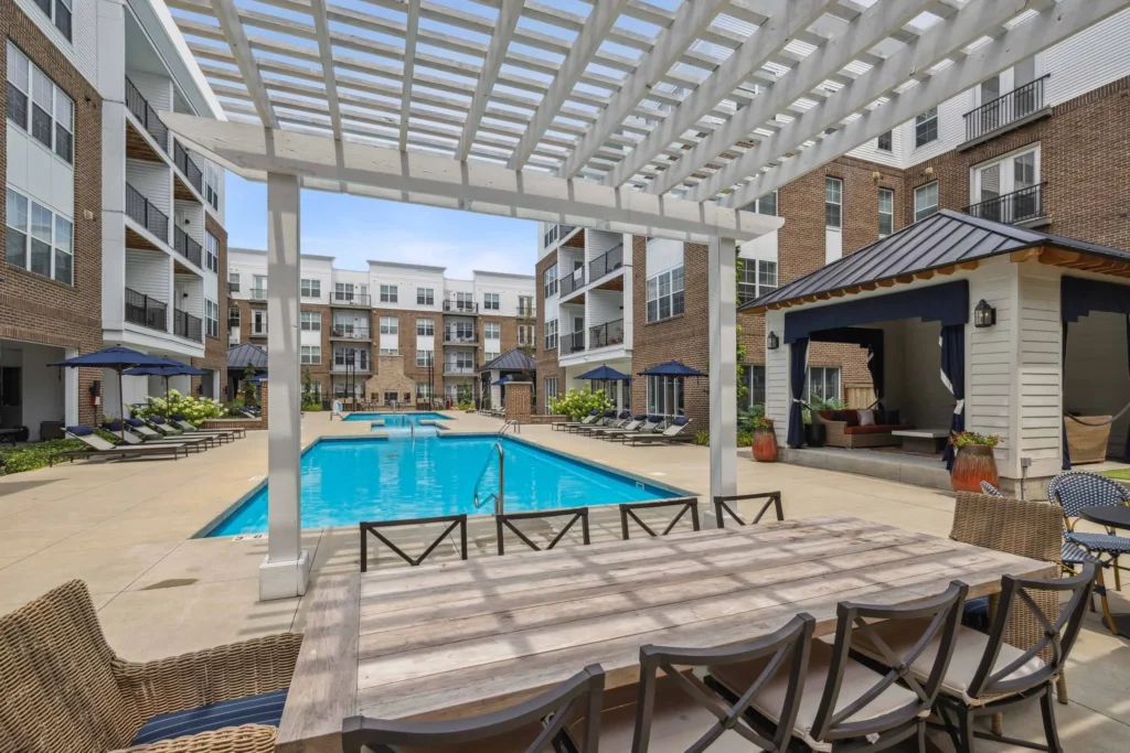 Outdoor courtyard with a swimming pool, lounge chairs, shaded cabanas, and a wooden dining table with chairs in a modern apartment complex.