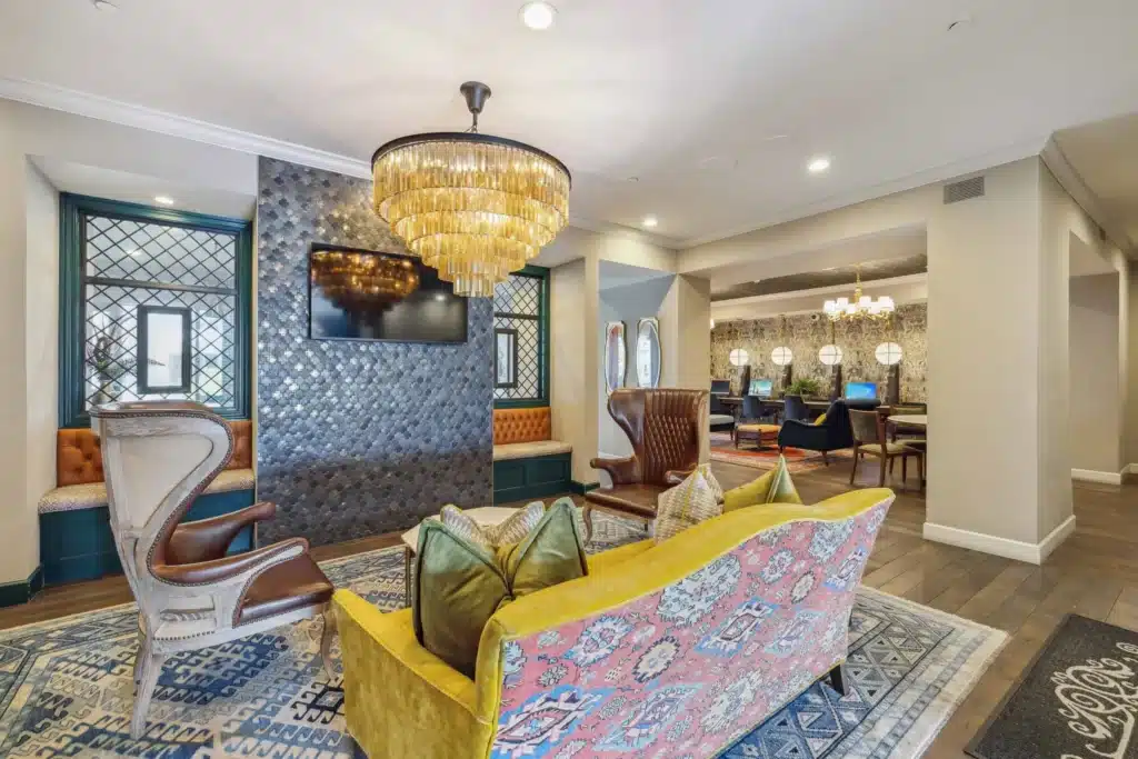 A modern lounge area with a yellow patterned sofa, high-back chairs, a wall-mounted TV, chandeliers, and a patterned accent wall; additional seating is visible in the background.