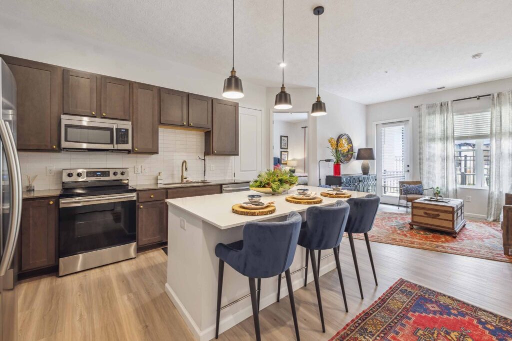 Modern kitchen with dark wood cabinets, stainless steel appliances, white island with three blue chairs, and an open living area with large windows and colorful rugs.