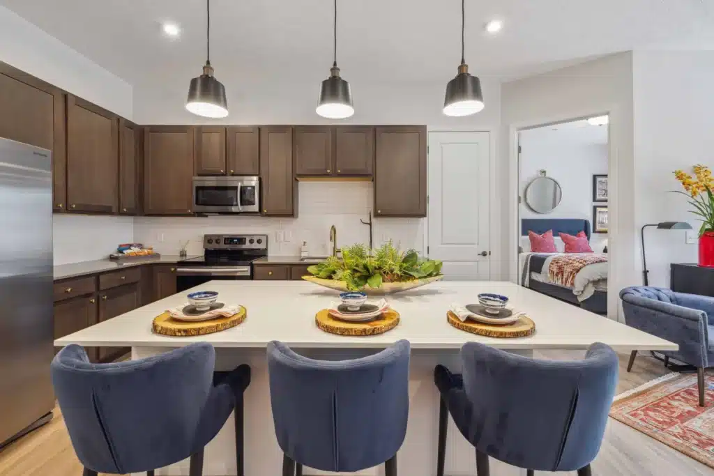 Modern kitchen with dark cabinets, stainless steel appliances, and an island set with three blue chairs and place settings; bedroom visible through open door.