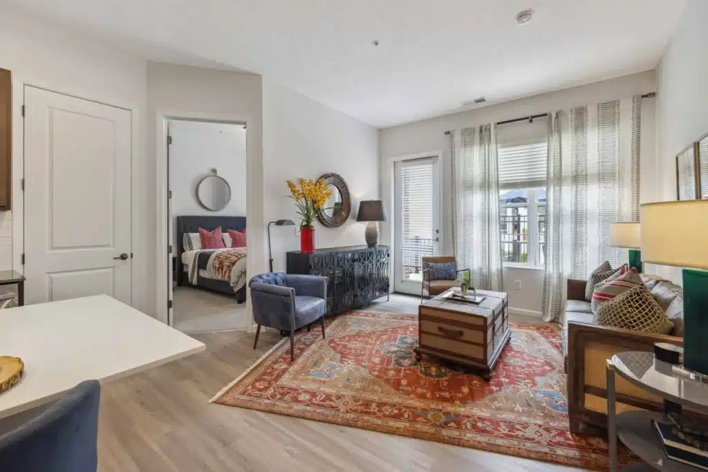 A modern living room with a red patterned rug, armchairs, a trunk coffee table, and large windows; a bedroom is visible through an open door.