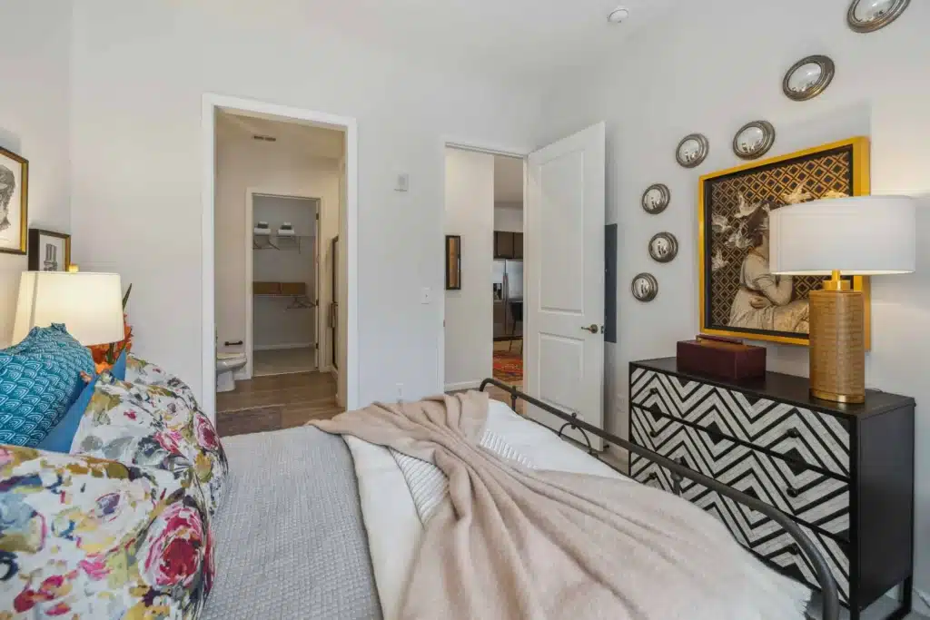 A neatly made bed with patterned pillows and a beige blanket in a bedroom featuring wall art, a dresser, a lamp, and an open door leading to a bathroom.