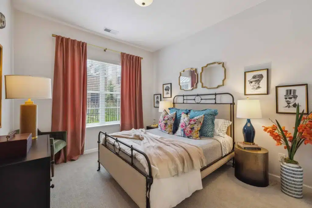 Bedroom with an iron bed frame, colorful pillows, two lamps, floral arrangement, artwork on the walls, and a window with red curtains letting in natural light.