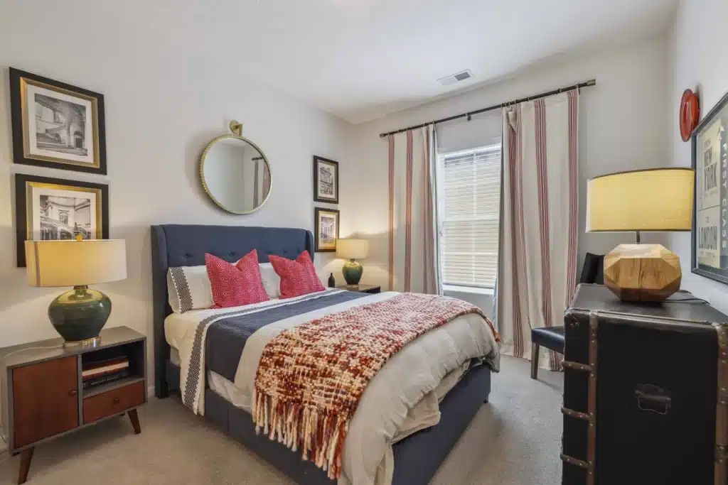 A neatly made bed with red accent pillows, a plaid throw blanket, two bedside tables with lamps, a dresser, and framed art on white walls in a bright bedroom.