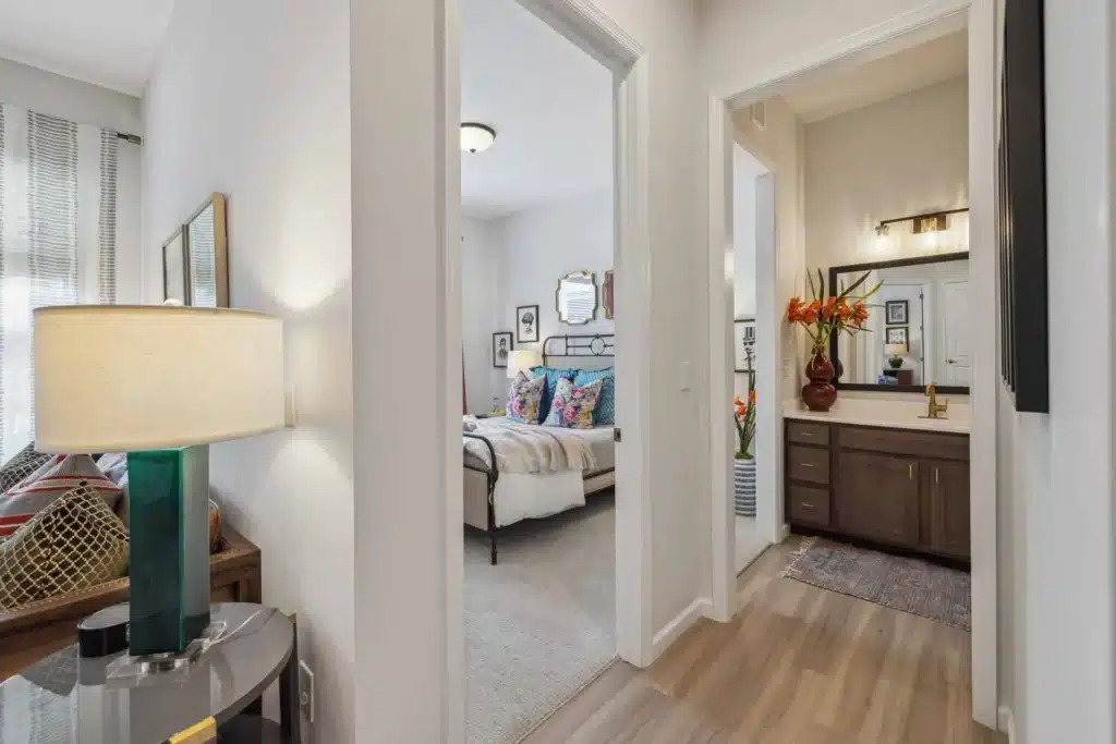 View down a hallway showing a bedroom with a metal bed frame and colorful pillows, and a bathroom with a dark wood vanity and orange flowers in a vase.