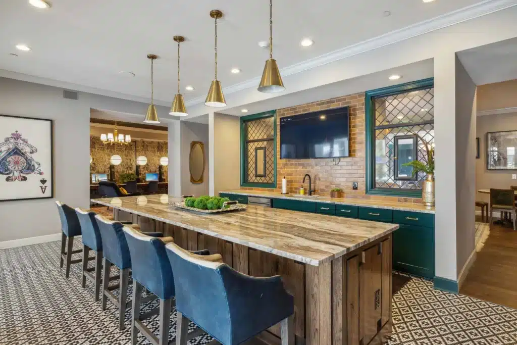 Modern kitchen with patterned floor, large marble island, navy upholstered bar stools, gold pendant lights, green cabinets, wall-mounted TV, and brick backsplash.
