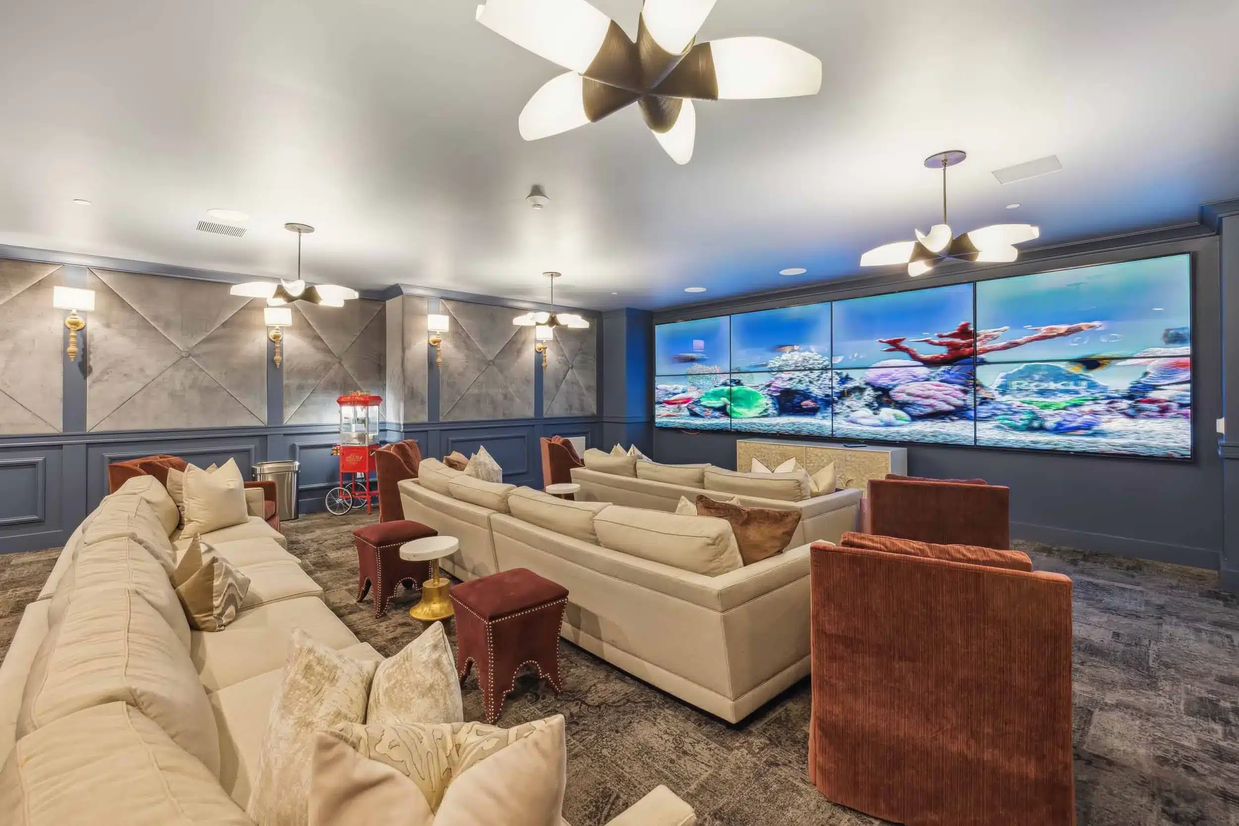 Home theater room with beige sofas, red armchairs, a popcorn machine, and a large screen displaying an underwater scene. The room features modern lighting and decorative wall panels.