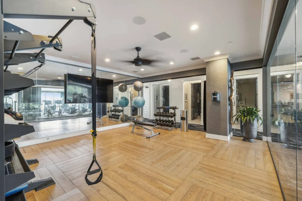 Modern home gym with exercise equipment, free weights, stability balls, a wall-mounted TV, mirrors, wooden flooring, and glass walls separating rooms.