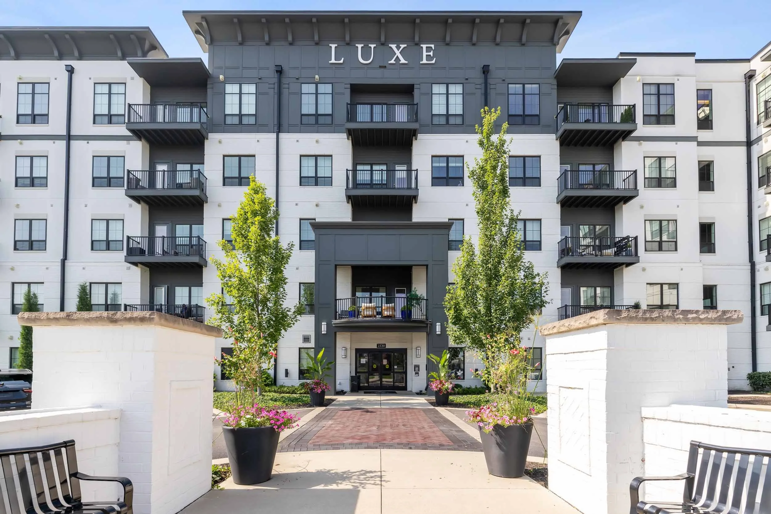 Modern apartment building with balconies and the word "LUXE" displayed at the top; landscaped entryway with planters and benches in the foreground.