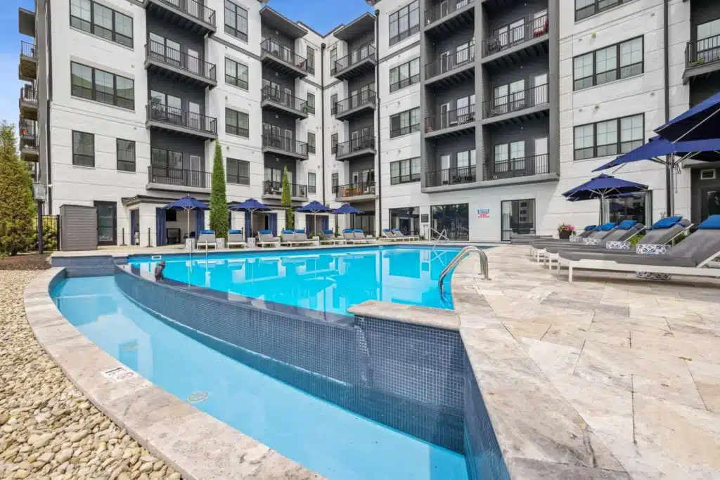 Outdoor swimming pool with lounge chairs and blue umbrellas, surrounded by a modern multi-story apartment building.