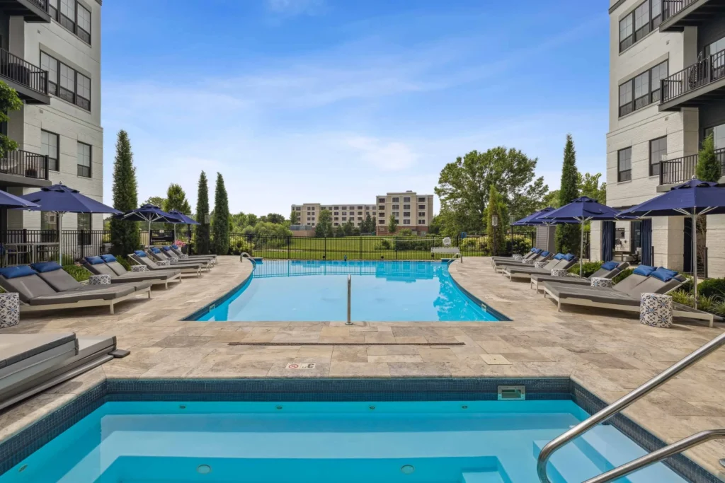 Outdoor swimming pool area with lounge chairs, umbrellas, and a hot tub, surrounded by modern apartment buildings and landscaped greenery.
